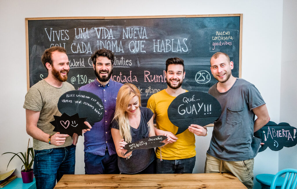 Four students standing in a classroom, in front of a board, smiling and holding up sheets with spanish phrases